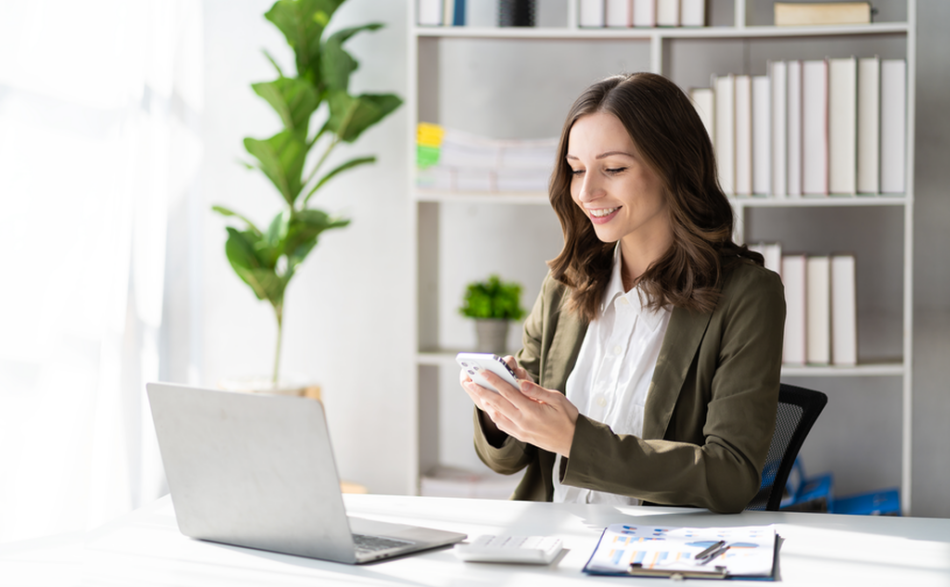 Kobieta w biurze, siedzi przy biurku, w rękach trzyma telefon komórkowy. Na biurku leży laptop i dokumenty. Wystrój pomieszczenia utrzymany jest w jasnej tonacji, w tle znajduje się regał na dokumenty oraz wysoki kwiat doniczkowy.