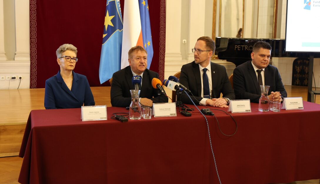 Na fotografii widoczne są cztery osoby siedzące przy stole konferencyjnym pokrytym bordowym płótnem. Od lewej strony przy stole siedzą: Beata Jagoda Członkini Zarządu MPEC Tarnów, Tadeusz Sieńczak Prezes Zarządu MPEC Tarnów, Jakub Kwaśny Prezydent Miasta Tarnowa oraz Maciej Włodek Zastępca Prezydenta Tarnowa. Fotografia została wykonana w Sali Lustrzanej w Tarnowie podczas konferencji prasowej. W tle widoczne są flagi Unii Europejskiej, Rzeczpospolitej Polskiej oraz Miasta Tarnów.