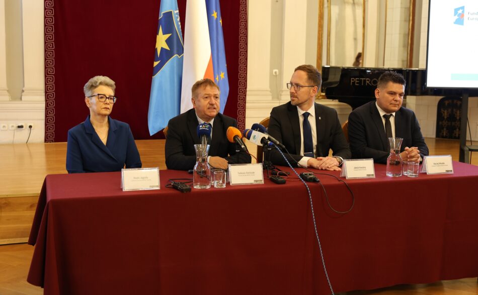 Na fotografii widoczne są cztery osoby siedzące przy stole konferencyjnym pokrytym bordowym płótnem. Od lewej strony przy stole siedzą: Beata Jagoda Członkini Zarządu MPEC Tarnów, Tadeusz Sieńczak Prezes Zarządu MPEC Tarnów, Jakub Kwaśny Prezydent Miasta Tarnowa oraz Maciej Włodek Zastępca Prezydenta Tarnowa. Fotografia została wykonana w Sali Lustrzanej w Tarnowie podczas konferencji prasowej. W tle widoczne są flagi Unii Europejskiej, Rzeczpospolitej Polskiej oraz Miasta Tarnów.