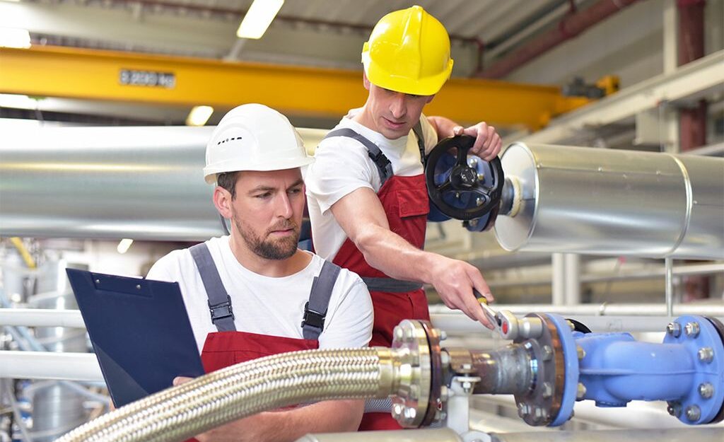 Na zdjęciu znajduje się dwóch mężczyzn w kaskach BHP. Pracują w pomieszczeniu technologicznym, wokół nich rozprowadzone są rury w kolorze srebrnym. Pierwszy z mężczyzn, w kasku w kolorze białym trzyma w dłoniach tablet, drugi, w kasku w kolorze żółtym trzyma dłoń na czarnym, dość dużym zaworze.