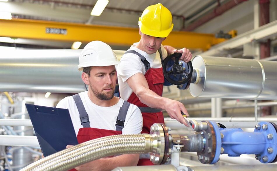 Na zdjęciu znajduje się dw&oacute;ch mężczyzn w kaskach BHP. Pracują w pomieszczeniu technologicznym, wok&oacute;ł nich rozprowadzone są rury w kolorze srebrnym. Pierwszy z mężczyzn, w kasku w kolorze białym trzyma w dłoniach tablet, drugi, w kasku w kolorze ż&oacute;łtym trzyma dłoń na czarnym, dość dużym zaworze.